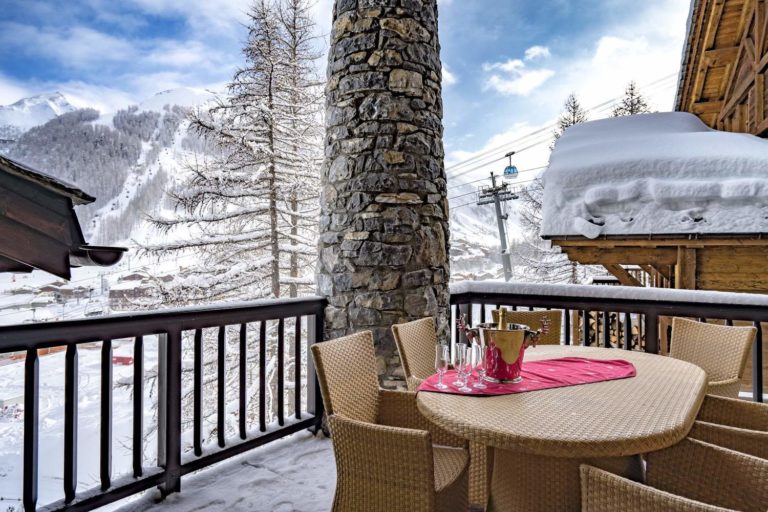 Terrasse en bois avec table ronde en osier et chaises, vue sur la montagne enneigée à Val d'Isère.